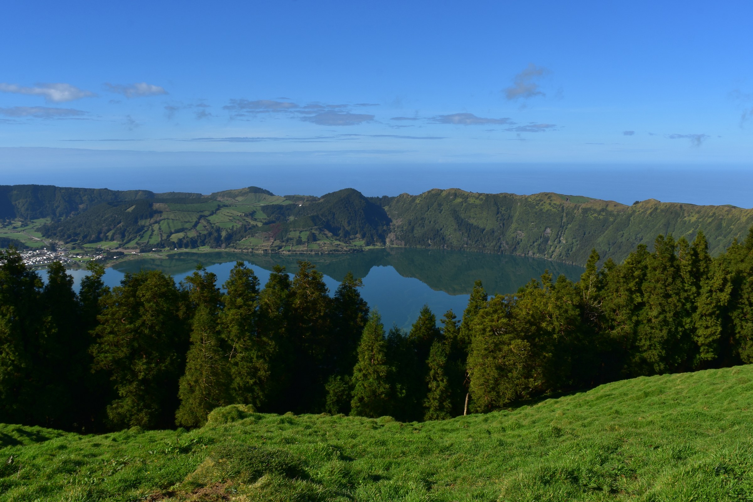 A still blue lake inside a ring of green hills, with dark pine trees and a grassy slope in front