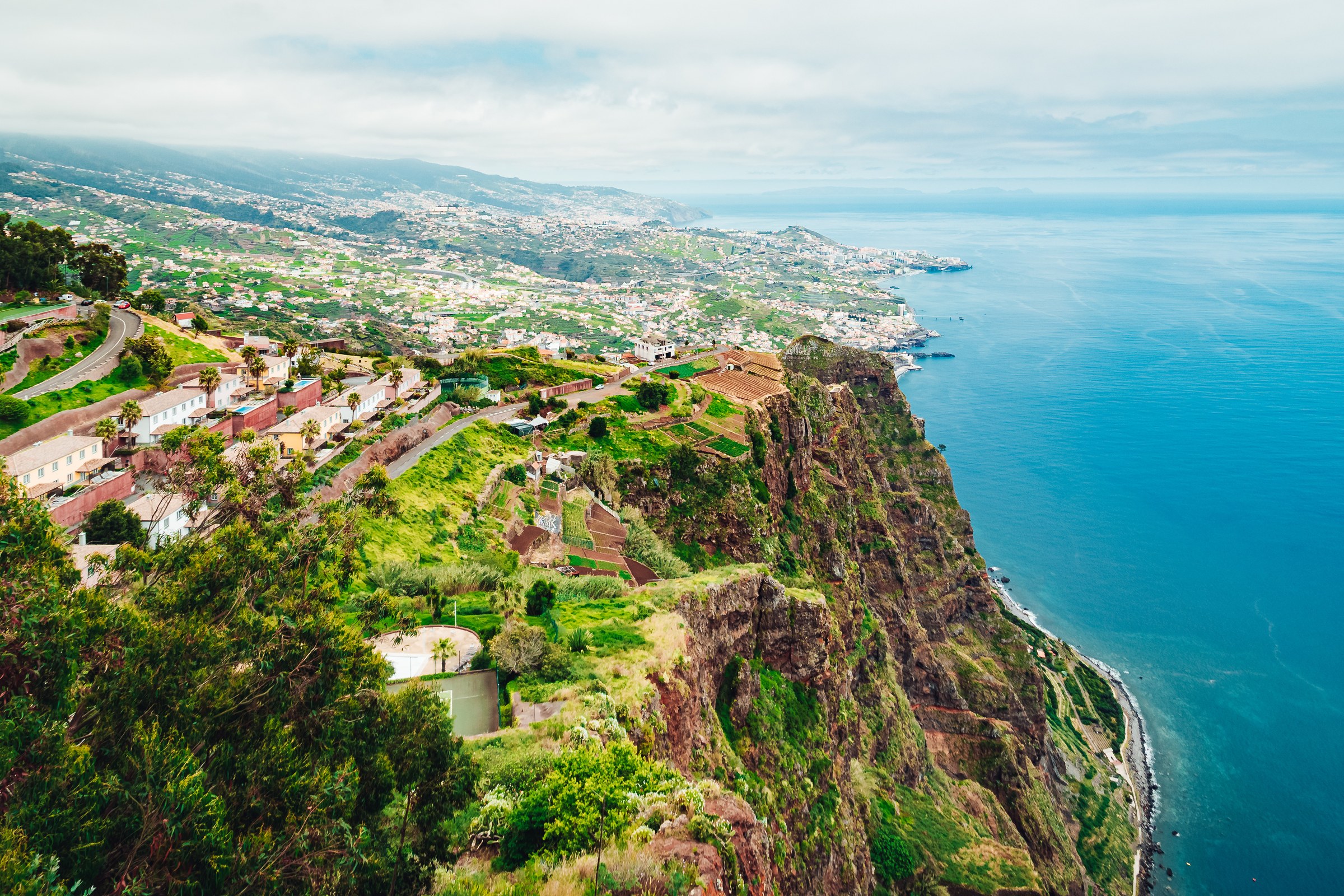A towering green sea cliff with terraced fields at its foot, beside a turquoise sea and a coastal town