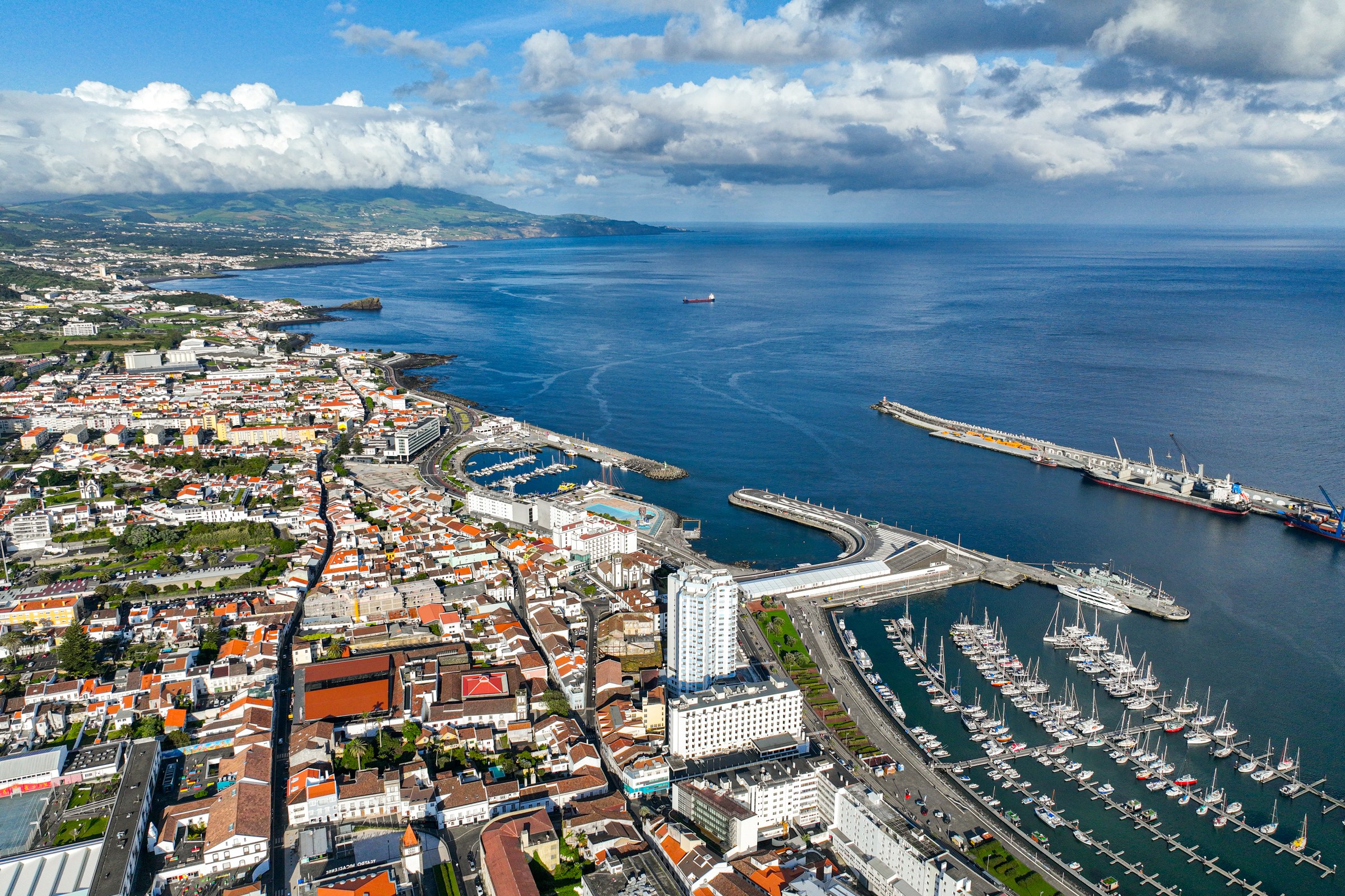 Aerial view of a white town beside a harbour with a long breakwater, a marina and a large ship docked