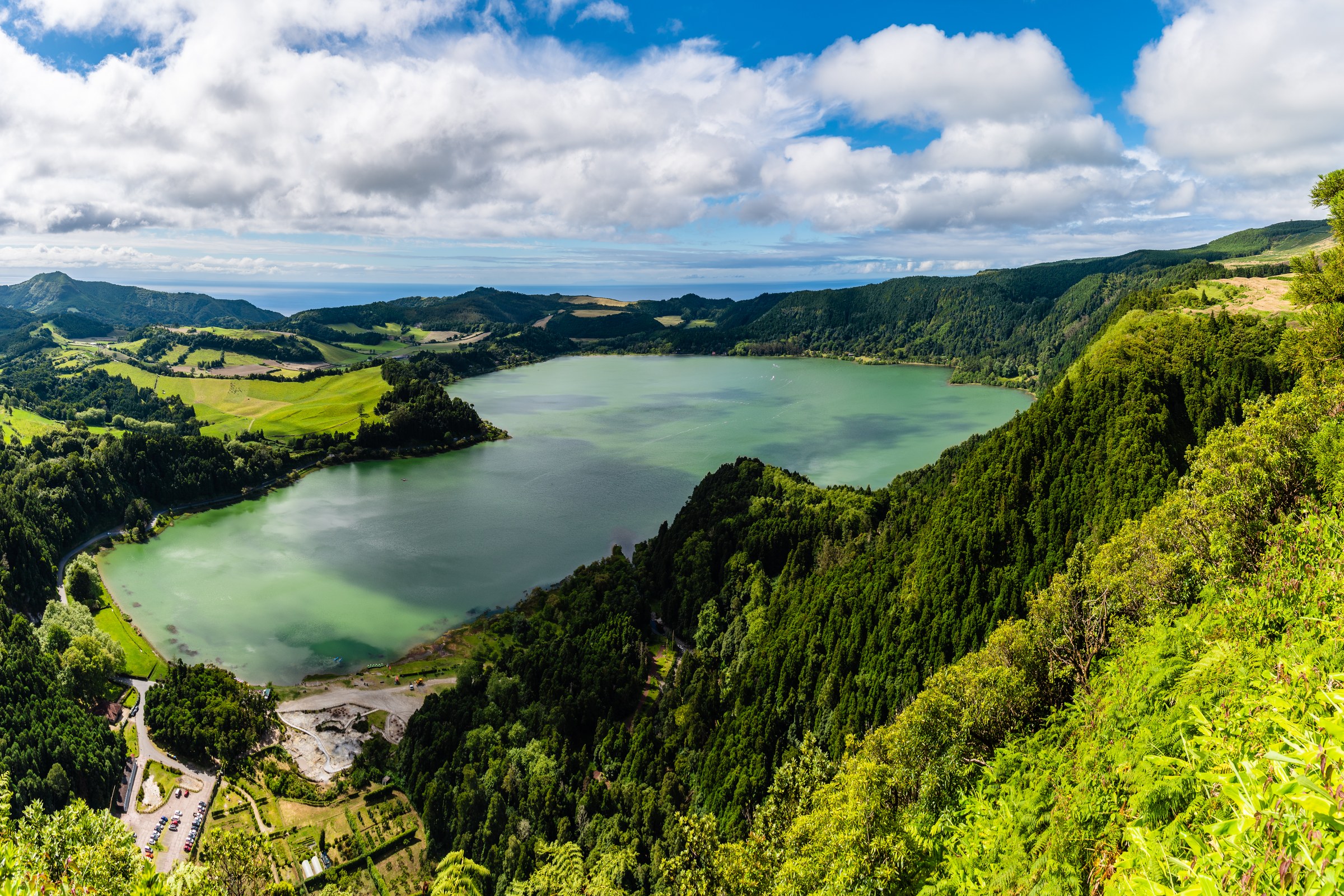 A pale green lake in a valley of steep wooded slopes and fields, under a blue sky with white clouds